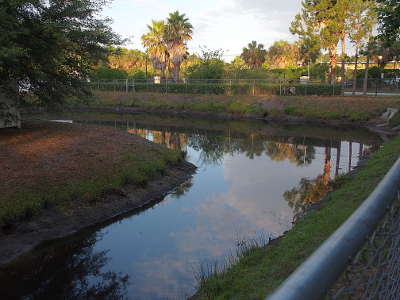 [This is an L-shaped pond with blue sky and clouds reflected in it. Based on the exposed dirt on the hillside, the water level is more than a foot low.]